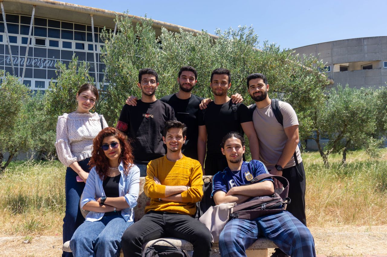 A diverse group of young adults posing together with smiles on a sunny day outdoors. University campus setting.